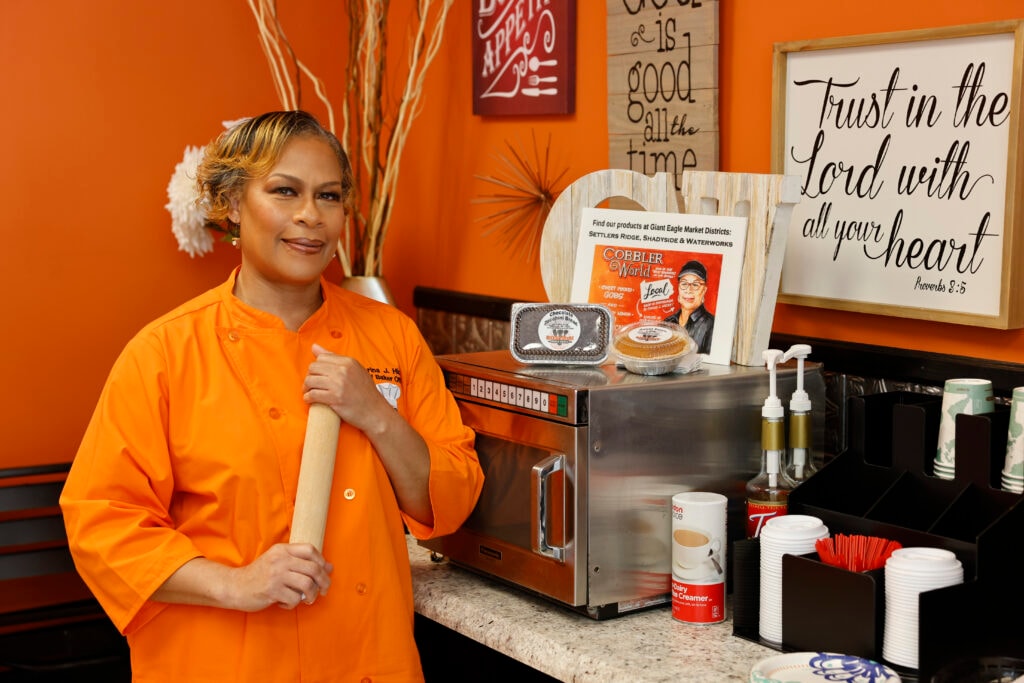 Terina J., owner of Cobbler World, in her orange chef coat holding a rolling pin