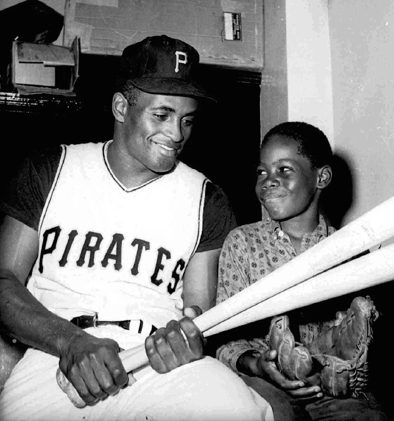 Roberto Clemente in his Pirates uniform sitting with a young fan holding bats