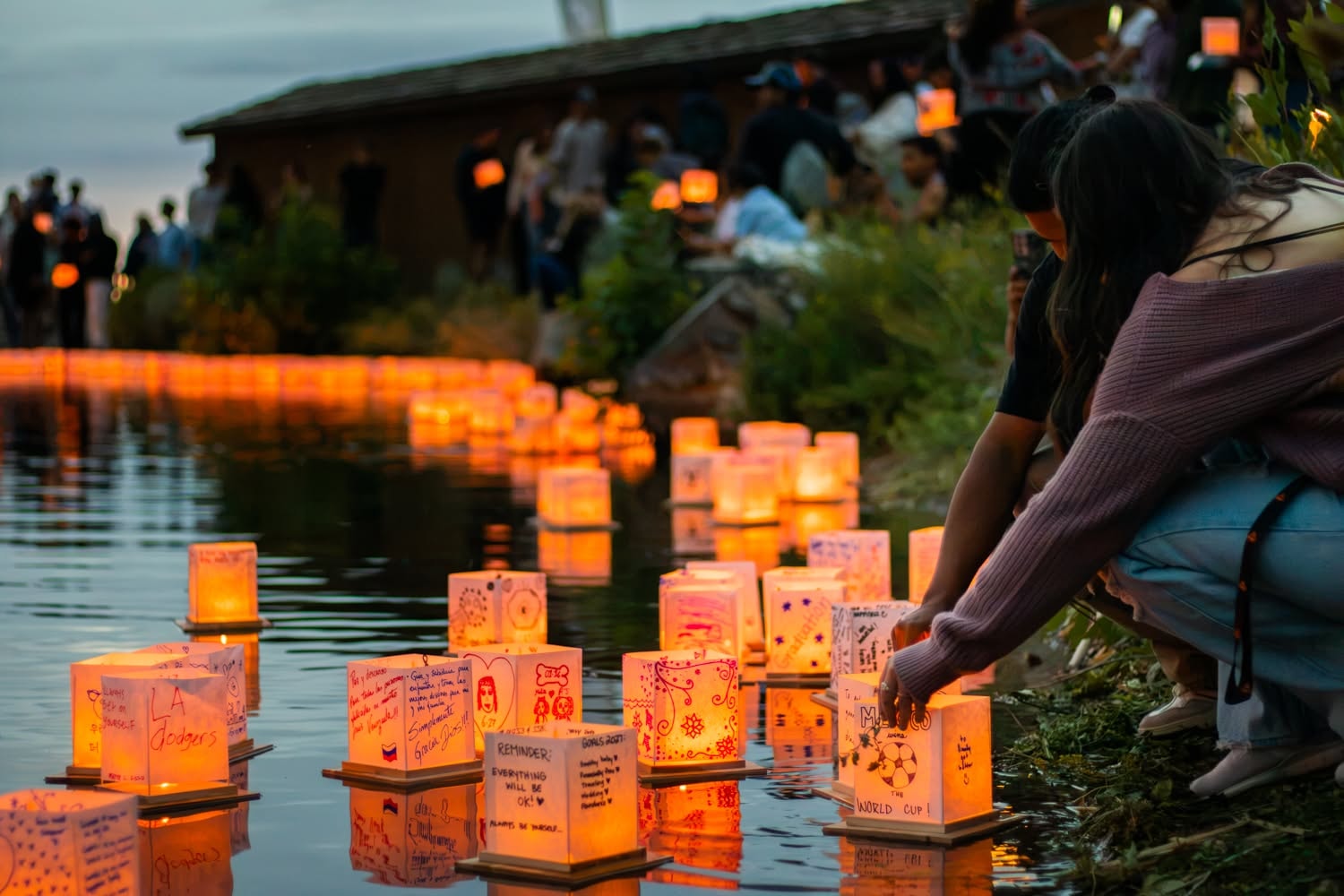 A woman kneels at the water's edge releasing a glowing lantern as crowds gather behind her